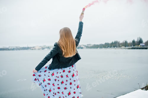 Preview: Stylish girl in leather jacket hold pink smoke flare at winter day against frozen lake.