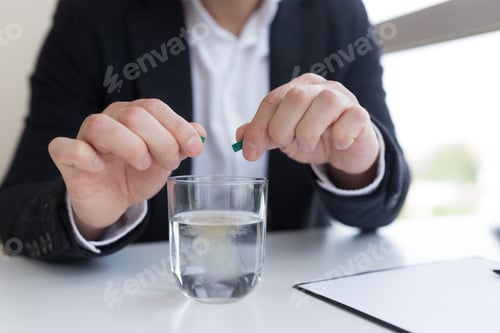 Preview: Close up of businessman in the office holding pills and glass of fresh water, taking medicine for