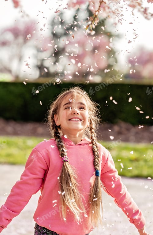 Preview: Happy excited little girl enjoying falling petals rain from blooming Sakura in park outdoors