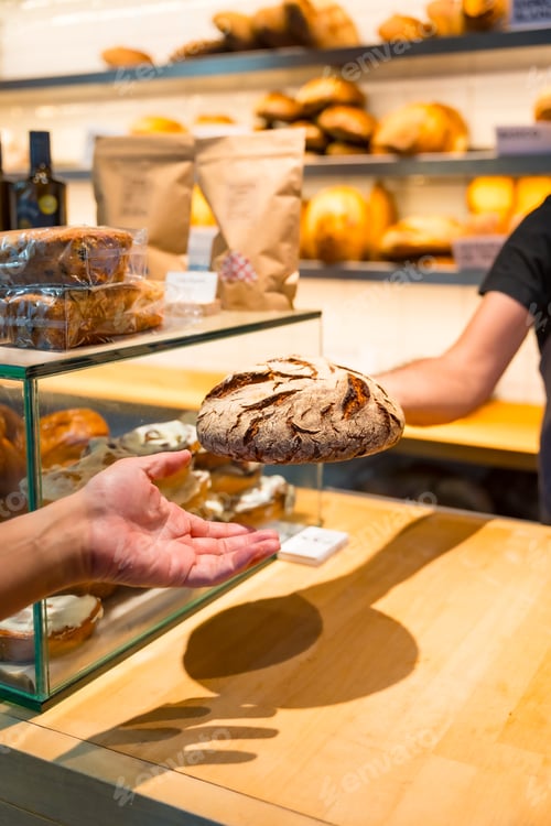 Preview: Hands of customer in the bakery shop buying an artisan bread from the baker in the workshop