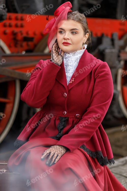 Preview: A beautiful girl in a burgundy suit is sitting on a suitcase near an old steam locomotive