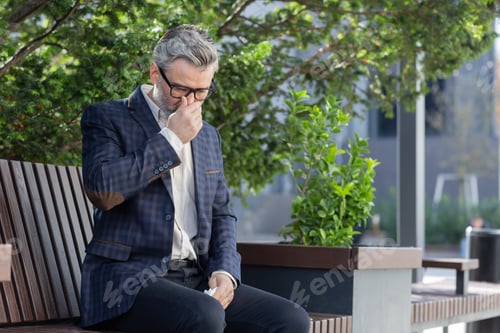 Preview: Sick and cold mature adult man sitting on bench outside office building, businessman in business