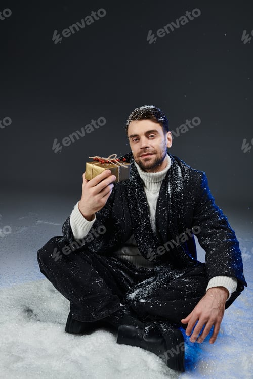 Preview: Handsome young man seated in snow, holding a beautifully wrapped gift with joy during wintertime
