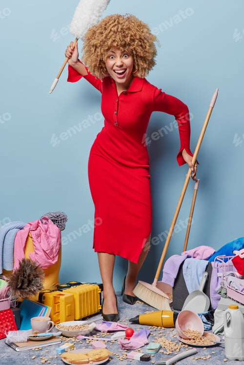 Preview: Full length shot of busy curly haired woman wears red dress holds dust brush and mop diligently