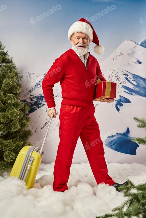 Preview: stylish Santa with white beard holding present in hand posing with yellow suitcase, winter concept