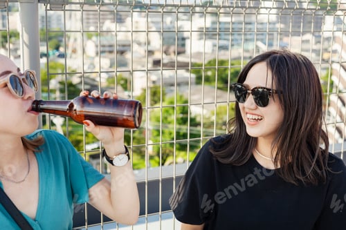 Preview: Two young Japanese women sitting on a rooftop in an urban setting, drinking beer.