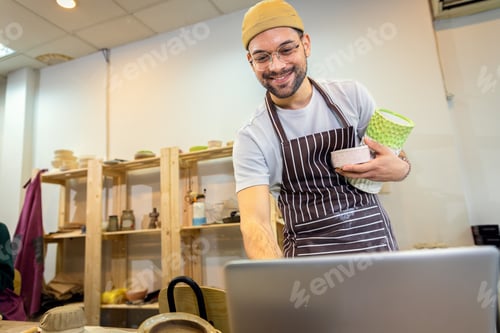 Preview: Male business owner using laptop in pottery studio workshop.