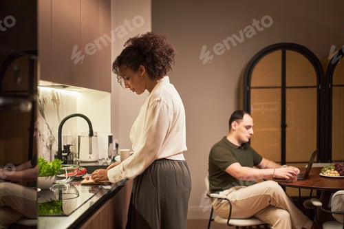 Preview: Young Adult Caucasian Man Using Laptop While Young Adult Black Woman Cooking