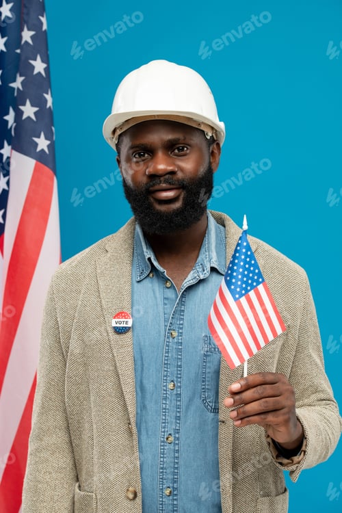 Preview: Young African builder or engineer in helmet and formalwear holding small us flag