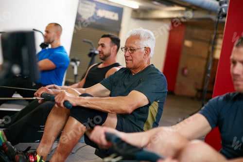 Preview: Cropped shot of a group of men working out on the rowing machine at the gym