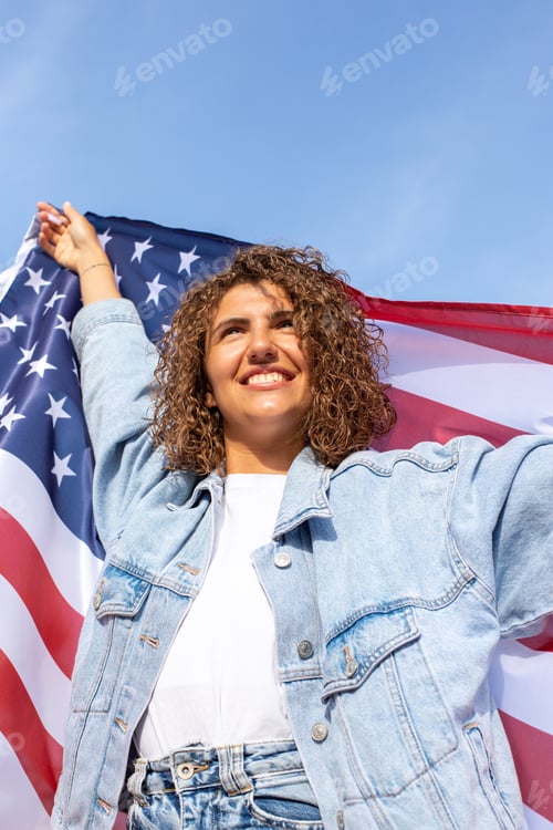 Preview: Beautiful woman in denim jacket with us flag celebrating Independence Day of America over blue sky