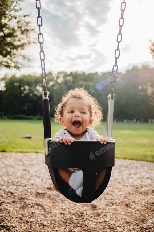 Preview: Cute curly hair mixed race baby boy on a swing in the park on a sunny summer day enjoying himself