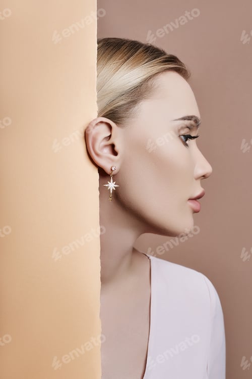 Preview: Earrings and jewelry in ear of a sexy blonde woman pressed against the paper beige