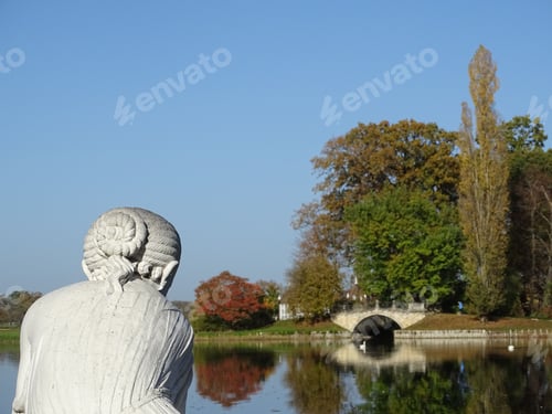 Preview: statue stands tall near a picturesque pond, with a bridge and autumn in the back in Woerlitz park
