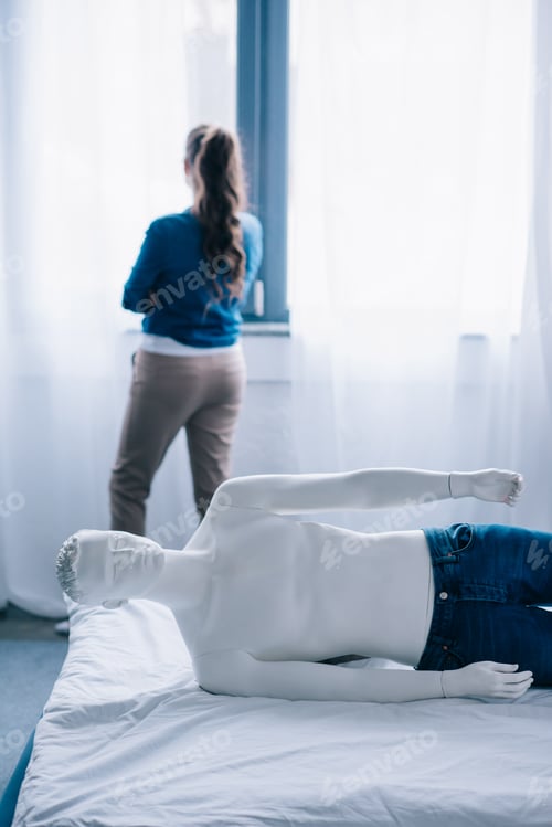 Preview: back view of woman standing at window and layman doll lying on bed, loneliness concept