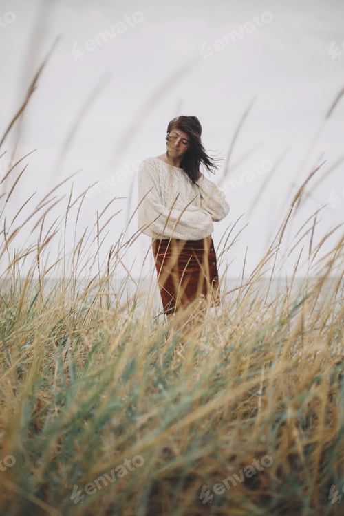 Preview: Beautiful stylish woman with windy hair in sweater posing among wild grass. Carefree moment.Tranquil