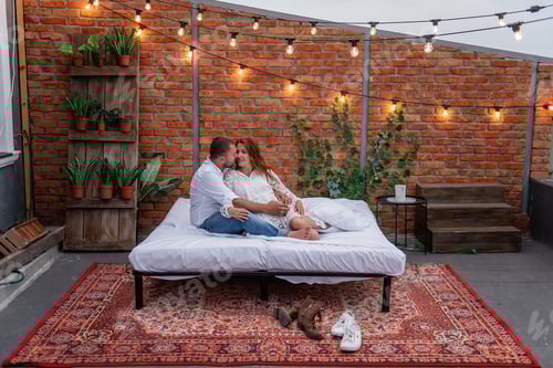 Preview: Loving couple millennials hug each other, resting on white bed. Background of brick wall, green ivy