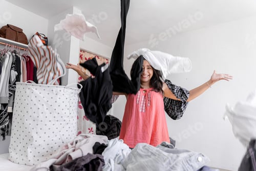 Preview: An Hispanic young woman is falsely smiling surrounded by dirty clothes flying overhead at home