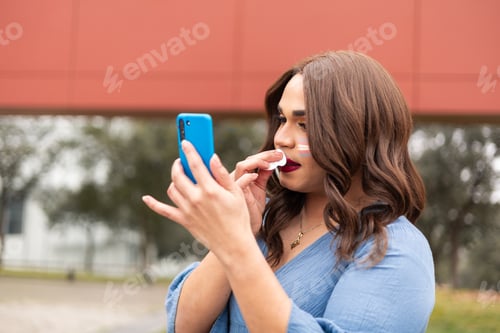 Preview: Young trans woman removing make up and using cellphone as mirror. LGTB lifestyle concept.