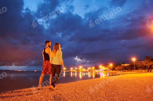 Preview: Man and woman resting on pebble beach at dusk on calm water dramatic blue cloudy sky background