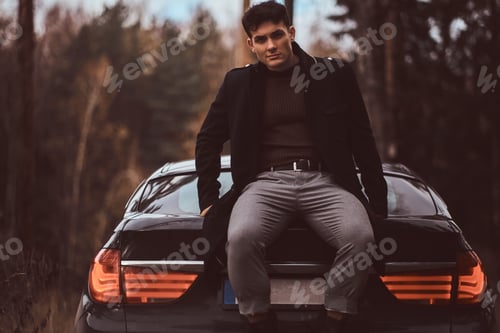 Preview: Stylish young man wearing a black coat standing next to a luxury car in the autumn forest