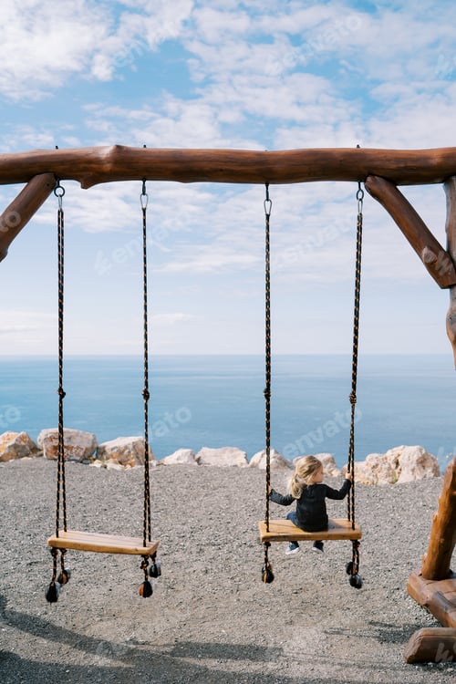 Preview: Little girl sits on a large wooden swing on a steep seashore and looks to the side. Back view