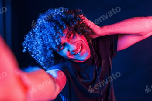 Preview: Takes selfie. Young beautiful man with curly hair is indoors in the studio with neon lighting