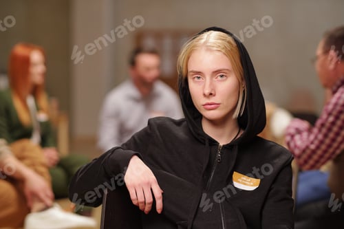Preview: Serious young blond woman in casualwear looking at you while sitting against mental support group