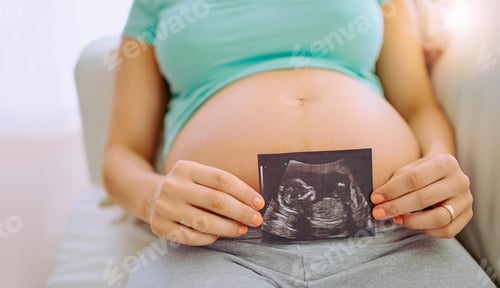 Preview: Closeup shot of a pregnant woman holding a sonogram picture against her belly at home
