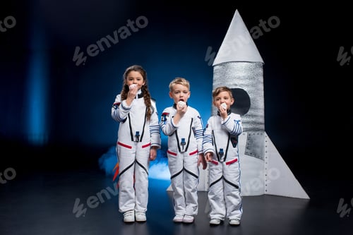 Preview: Three cute kids in astronaut costumes eating sweets standing near rocket