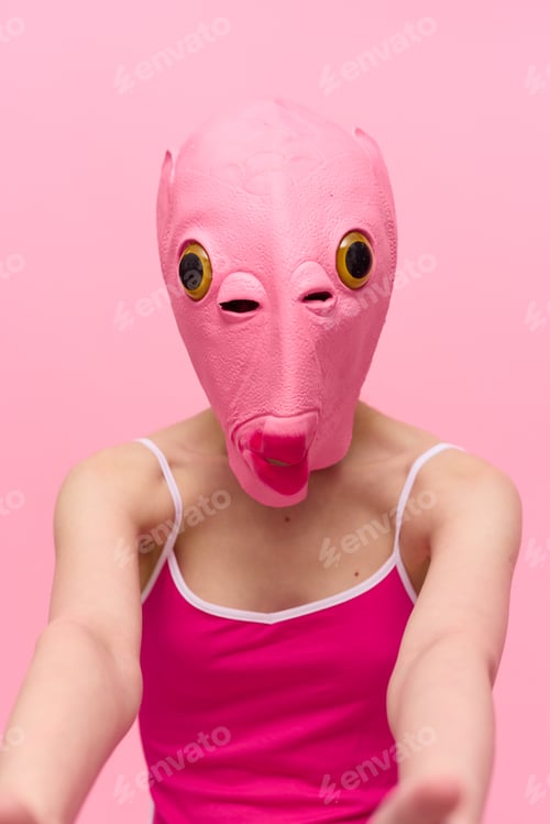 Preview: A woman wearing a silicone Halloween mask in the shape of a pink fish with big yellow eyes looks at