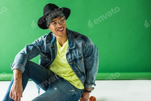 Preview: stylish mixed race man sitting, smiling and posing on green with copy space