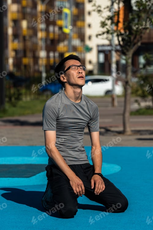 Preview: Adult asian man in glasses sitting exhausted after doing sports in park.