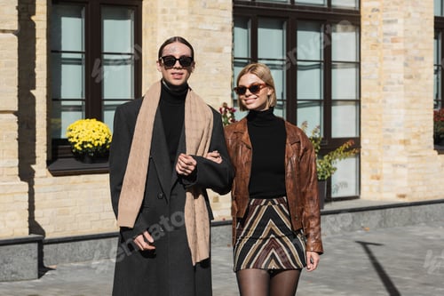 Preview: tattooed man in sunglasses and coat standing with happy blonde woman in leather jacket
