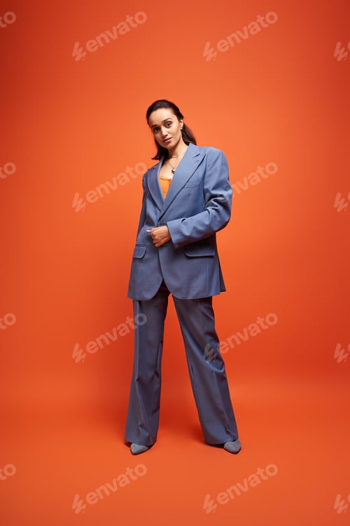 Preview: Brunette model showcases trendy blue suit against vibrant orange backdrop