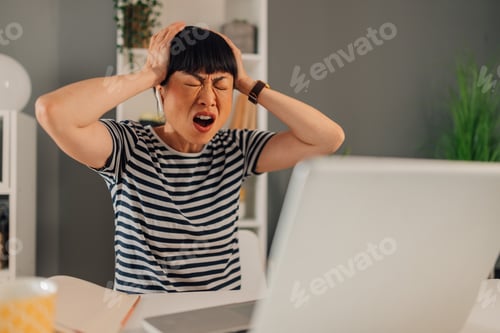 Preview: Asian woman holding head expressing frustration while working remotely