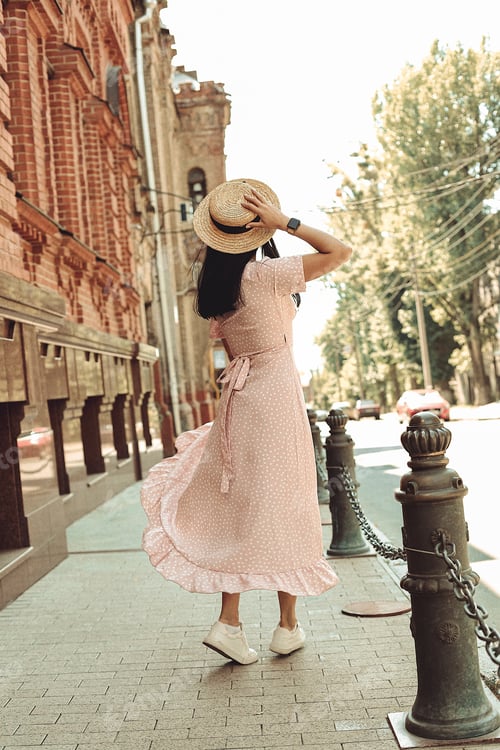 Preview: A young brunette in a soft pink dress with a hat walks around the city