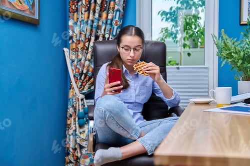 Preview: Teenage girl sitting on armchair near computer, looking in smartphone, eating snack