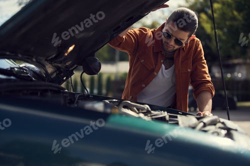 Preview: pensive stylish male model with ponytail and trendy accessories looking at engine of his car