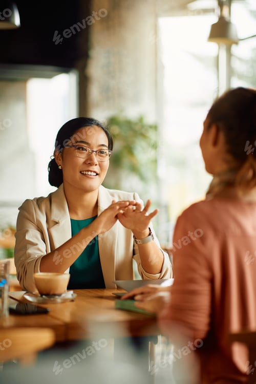 Preview: Happy Asian entrepreneur talking to her colleague during coffee break in a cafe.