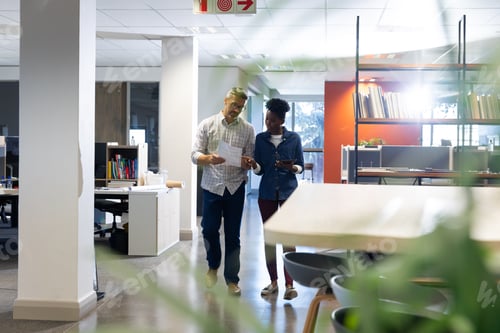 Preview: Biracial colleagues discussing over document while walking together at modern workplace