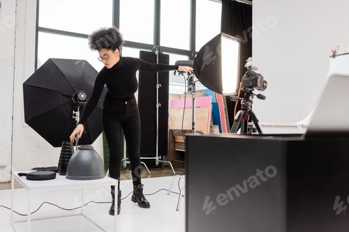Preview: african american content producer in black clothes reaching lamps near softbox reflector and digital