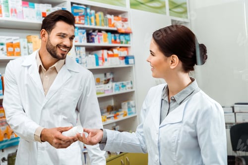 Preview: Smiling pharmacist giving to colleague jar with pills in pharmacy