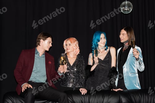 Preview: smiling queer friends with champagne glasses talking during new year party near leather couch on