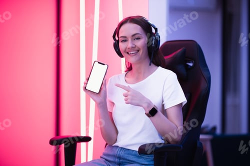 Preview: Cheerful gamer girl pointing with finger at smartphone with blank screen while sitting on a gaming