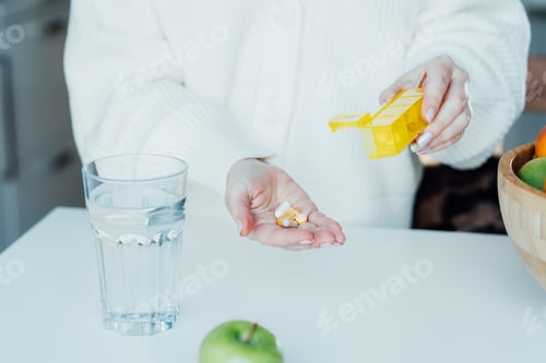 Preview: Close-up of woman's hand holding pills and pill organizer at home kitchen with water glass.