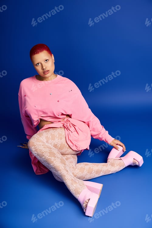 Preview: Young woman with short pink hair poses boldly in a studio against a vibrant blue backdrop