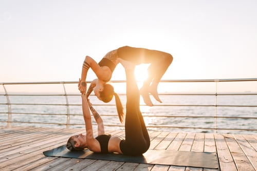 Preview: Photo of two young ladies in black sporty tops and leggings training yoga poses by the sea
