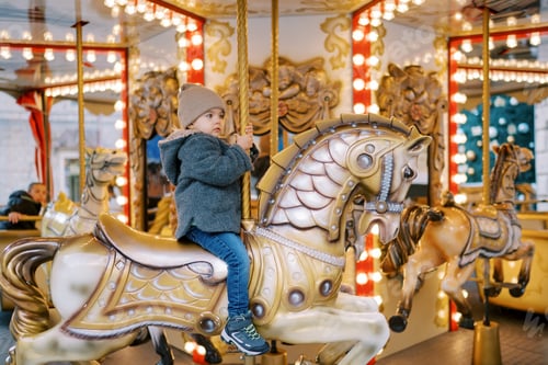 Preview: Little girl sits on a toy horse on a carousel, holding the handle. Side view