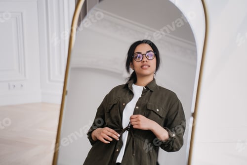 Preview: Young African American lady in casual shirt standing at mirror at room looks away with pensive face